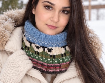 May include: A knitted cowl neck warmer featuring a blue ribbed collar, a pattern of white sheep on a green background, and horizontal stripes in pink, white, and dark gray. A small rectangular brown leather tag is attached to the lower edge.