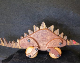 May include: Wooden toy dinosaur with brown and yellow striped wood, four wheels, and red accents. The dinosaur has a long tail and spikes on its back.