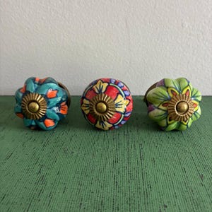 May include: Three decorative ceramic cabinet knobs. One is teal with orange accents, another is red and yellow, and the third is green and yellow. Each knob has a brass-colored base and a floral design.