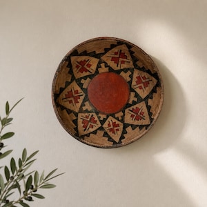 May include: A round, shallow wooden bowl featuring a central solid red circle surrounded by a repeating geometric pattern of black and red diamond shapes on a natural tan wood background, creating a star-like design across the interior surface.