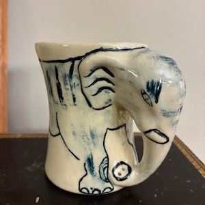 May include: A ceramic mug with a whimsical elephant design. The mug is off-white with blue painted details, including the elephant's outline and decorative stripes. The handle is shaped like the elephant's trunk. A unique and artistic piece.