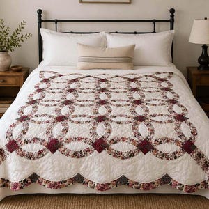 May include: A white quilt featuring a double wedding ring pattern with floral fabric in shades of burgundy, pink, and green. The quilt has a scalloped edge and is draped over a bed with white pillows and a rectangular lumbar pillow with stripes.