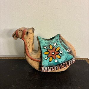May include: A ceramic camel figurine with a turquoise saddle decorated with a yellow and orange floral design. The camel is tan with red accents on the head and legs. The figurine is resting on a dark surface.