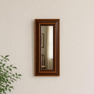 May include: A rectangular wall mirror featuring a deep brown wooden frame with a multi-layered, stepped profile. The glass surface reflects a portion of a room, including a wooden chair and a window with light-colored curtains in the background.