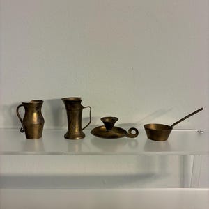 May include: A collection of small, antique brass items displayed on a clear shelf. The items include a small pitcher, a vase, a candle holder, and a small pan with a long handle. The objects have a warm, golden color.