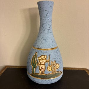 May include: A light blue ceramic vase with a speckled texture. The vase features a gold band and a painted scene of a person with a donkey and cactus. The vase is approximately 8 inches tall.