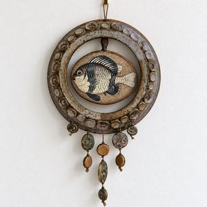 May include: A circular ceramic wall hanging featuring a central oval plaque with a detailed illustration of a fish. The outer ring is decorated with small, raised circular elements. Below the ring, several ceramic beads hang on thin cords.