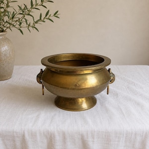 May include: A polished brass bowl with a wide, circular rim and a pedestal base. The bowl features two decorative side handles, each adorned with a small, dangling ring. The metal surface has a warm, golden tone with subtle variations in luster.