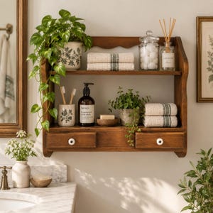 May include: A two-tiered wooden wall shelf with two drawers featuring white knobs. The shelf holds a potted trailing plant, a glass jar with cotton balls, a reed diffuser, folded textured towels, a toothbrush holder, and a hand wash bottle.
