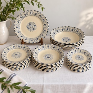 May include: A collection of cream-colored ceramic dinnerware featuring a blue floral pattern with a central motif and a decorative border. The set includes multiple stacked dinner plates, soup bowls, and smaller side plates arranged on a table.