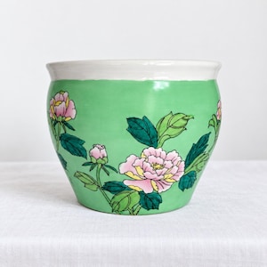 May include: A ceramic planter with a wide, white rim and a bright green body. The exterior features a hand-painted floral design of pink peonies with yellow centers, accompanied by dark green leaves and stems wrapping around the curved surface.