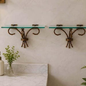 May include: Two wall-mounted shelves featuring clear glass rectangular surfaces supported by bronze-toned metal brackets. Each bracket has a twisted metal design with decorative scrollwork and a central knot detail, mounted to a light-colored wall.