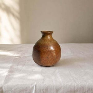May include: A small, round ceramic vase with a narrow neck and a wide, bulbous body. The surface features a mottled, earthy glaze with blended tones of deep brown, rust orange, and muted ochre, creating a textured, organic appearance throughout.
