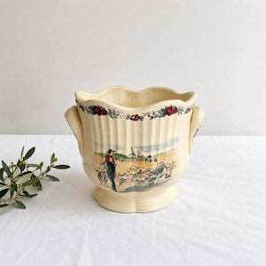 May include: A cream-colored ceramic planter with a scalloped rim and two side handles. The exterior features vertical ribbing and a colorful illustration of a person standing in a field with a windmill, topped by a floral border of red roses.