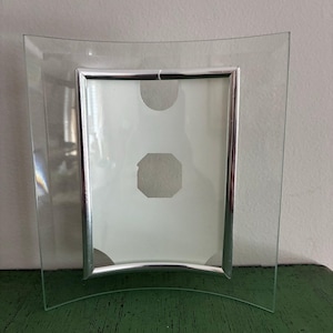 May include: Clear glass photo frame with a curved design. The frame has a silver metal border around the photo area. The back of the frame is white with two circular and one octagonal cutouts.