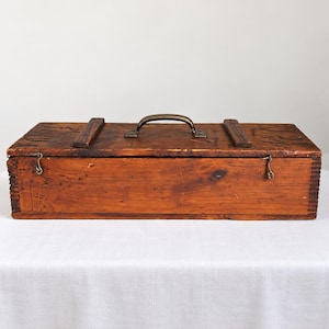 May include: A rectangular wooden storage box with a dark brown finish, featuring a metal handle on the top lid, two small metal hook latches on the front, and visible finger joint construction on the side edges of the box.