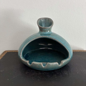 May include: A teal ceramic ashtray with a glossy finish, featuring a rounded, dome-like shape with a large front opening and a small, upright chimney-style vent at the top. The edges of the opening and the base show subtle brown-toned detailing.
