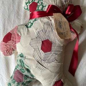 May include: A handmade patchwork teddy bear in a whimsical design. The bear features a variety of fabric patterns, including floral, striped, and solid red accents. A red ribbon is tied around the bear's neck, and a tag reads "Treasure Pie, take your own family heirloom, Hand Made in USA."