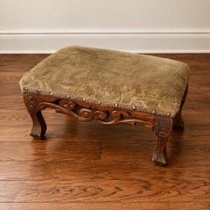 May include: A rectangular footstool featuring a weathered olive-green fabric top with a textured surface. The base is constructed from dark brown wood with intricate carved scrollwork details and four curved legs, accented by a row of metal nailheads.