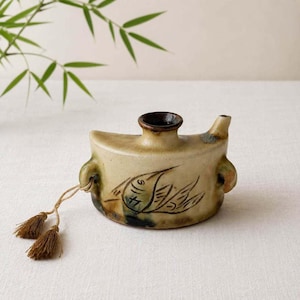 May include: A beige ceramic oil lamp with a dark brown rim and a spout. The body features a hand-painted black line drawing of a fish. Two small handles on the sides hold a thin cord with two small brown tassels attached to one side of the vessel.