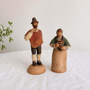 May include: Two hand-painted clay figurines standing on a surface. One figure wears a brown hat, a rust-colored poncho, and dark shorts. The other figure wears a green top and a tan skirt, holding a small brown pot with geometric patterns.
