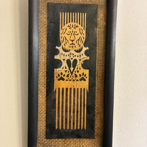 May include: Framed artwork featuring a wooden comb design with a lion's head and paw prints. The comb is gold-colored against a black background, framed in black with a woven texture. A decorative piece for home decor.