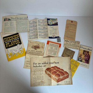 May include: A collection of vintage paper recipe booklets and pamphlets featuring various food illustrations and text including Chicken with a New Flavor, Tried Recipes, 23 New Dishes, and For an added treat next bakeday with a loaf of bread image.