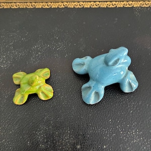 May include: Two ceramic frog figurines. One is a bright green with yellow accents, and the other is a light blue. Both frogs are in a sitting position with their legs spread out.