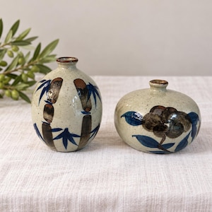May include: Two ceramic vases with a speckled off-white glaze. The taller, rounded vase features hand-painted brown bamboo stalks with blue leaves. The shorter, flattened vase displays a dark brown floral motif with blue leaves and stems.