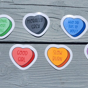 Könnte beinhalten: Acht herzförmige Aufkleber mit verschiedenen Sprüchen, darunter "Emotional Support Kindle", "Morally Grey", "Who Did This To You?", "Friends To Lovers", "Good Girl", "Slow Burn", "Enemies To Lovers".