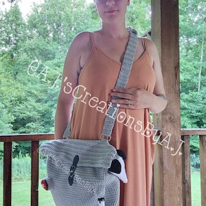 Op de afbeelding: Een vrouw in een perzikkleurige jurk houdt een gehaakte grijze tas vast met een wit en zwart gehaakt dier eraan bevestigd. De tas heeft een zwarte band en een zwarte gesp. De tekst "GAJ's CreationsByAJ" is zichtbaar op de tas.