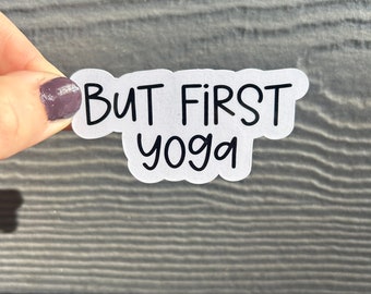 May include: A white sticker with the text "But First Yoga" in black, handwritten-style font. The sticker is held by a hand with purple nail polish against a gray wooden background. The sticker is a motivational item.