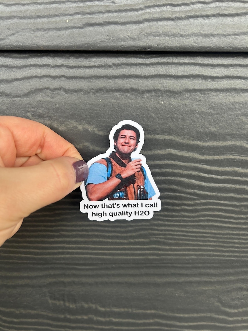 K&ouml;nnte beinhalten: Ein Aufkleber mit einem l&auml;chelnden Mann in blauem Hemd und Weste, der eine Tasse h&auml;lt. Der Aufkleber tr&auml;gt den Text "Now that's what I call high quality H2O". Der Aufkleber wird von einer Hand mit einem lila Nagel gehalten.