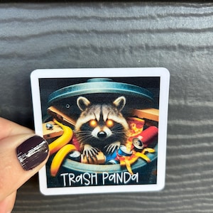 May include: A white sticker with a black background featuring a raccoon with glowing red eyes inside a trash can. The raccoon is surrounded by various items like banana peels, cans, and pizza crusts. The text "Trash Panda" is printed in white at the bottom of the sticker.