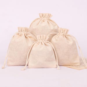 May include: A collection of five beige burlap drawstring bags of varying sizes. Each bag has a gathered top and a matching drawstring closure. The bags are arranged in a group, with the largest bag in the back and the smallest in the front.