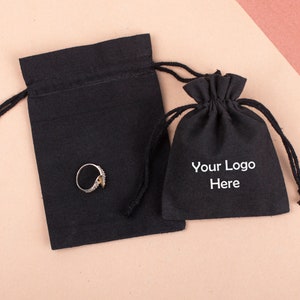 Op de afbeelding: Twee zwarte zakjes met trekkoord, een rechthoekige en een ronde, met een ring in het rechthoekige zakje. Het ronde zakje heeft de witte tekst "Your Logo Here". De ring is goudkleurig met kleine, heldere stenen.