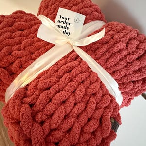 May include: A chunky knit blanket in a warm coral color, tied with a cream ribbon. A small card with the text "Your order made day" is attached. The blanket is folded and appears soft and cozy.