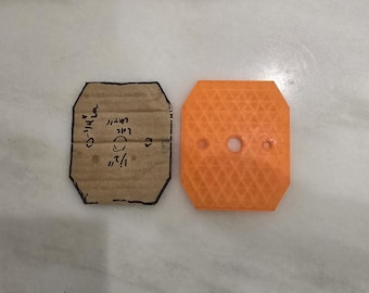 May include: A rectangular cardboard template with clipped corners and handwritten measurements of 1/2 inch and 1-1/4 inch next to an orange 3D-printed plastic part with a matching shape, a textured diamond pattern surface, and three circular holes.