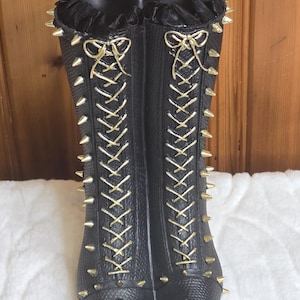 May include: A pair of black boots with a textured surface, featuring gold-toned lace-up fronts tied with bows at the top. The boots are adorned with rows of sharp gold spikes along the sides and have black ruffled trim around the top openings.