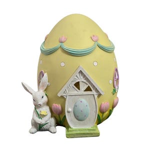 May include: A yellow egg-shaped house with a white door and a small bunny rabbit figurine standing in front. The house has a blue and green trim and pink flowers around the base.