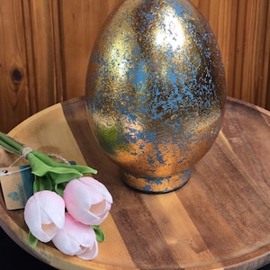 May include: A large, decorative egg with a gold and blue metallic finish sits on a wooden tray. The egg is paired with a small bouquet of pink tulips. The egg is approximately 10 inches tall, making it a striking Easter or spring decoration.