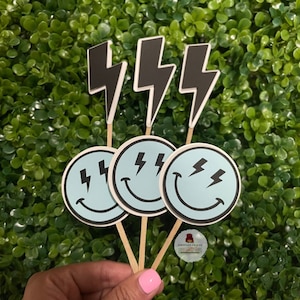May include: Three cupcake toppers with a light blue smiley face design and black lightning bolts. The toppers are on wooden sticks, with black lightning bolt shapes above the smiley faces. The background is a green hedge.