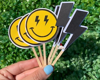 May include: A set of cupcake toppers featuring yellow smiley faces with lightning bolts and black lightning bolt shapes. The toppers are attached to wooden sticks.