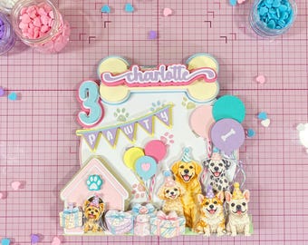 May include: A vibrant dog-themed cake topper for a third birthday, featuring the name "Charlotte". It includes various dog breeds, balloons, and the text "PAWTY". The background is pink with heart-shaped confetti scattered around the design.
