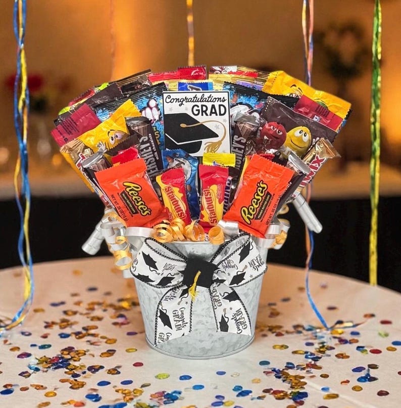 May include: A galvanized metal bucket filled with assorted candy bars and treats, arranged as a gift basket. The bucket is decorated with a graduation-themed bow and the words "Congratulations Grad". A card with a graduation cap is visible.