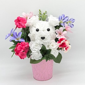 May include: A white dog-shaped flower arrangement in a pink pot. The arrangement is made of white roses and other flowers in shades of pink, purple, and white.