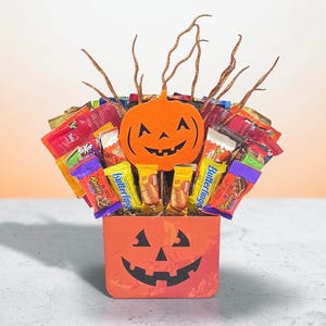 May include: A Halloween candy bouquet featuring an orange felt jack-o'-lantern cutout and glittery brown twigs. The arrangement is housed in an orange square container with a black jack-o'-lantern face, filled with assorted candy bars.