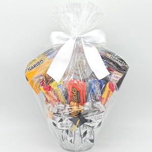 May include: A clear cellophane-wrapped gift basket filled with assorted candy bars and treats. The basket is tied with a white satin bow. The base of the basket is decorated with a graduation-themed design, including mortarboards and the words "Congrats Grad".