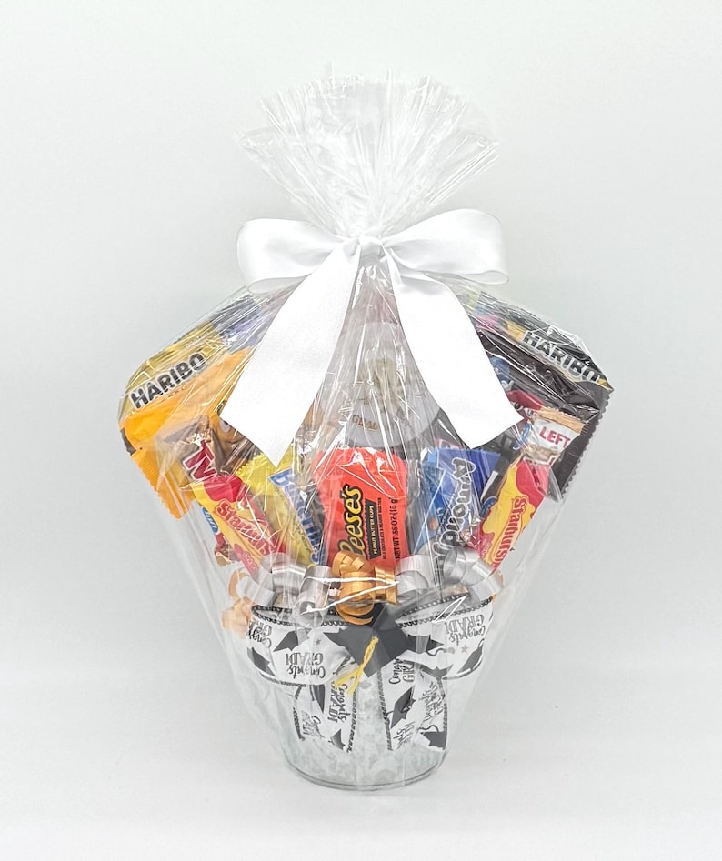 May include: A clear cellophane-wrapped gift basket filled with assorted candy bars and treats. The basket is tied with a white satin bow. The base of the basket is decorated with a graduation-themed design, including mortarboards and the words "Congrats Grad".
