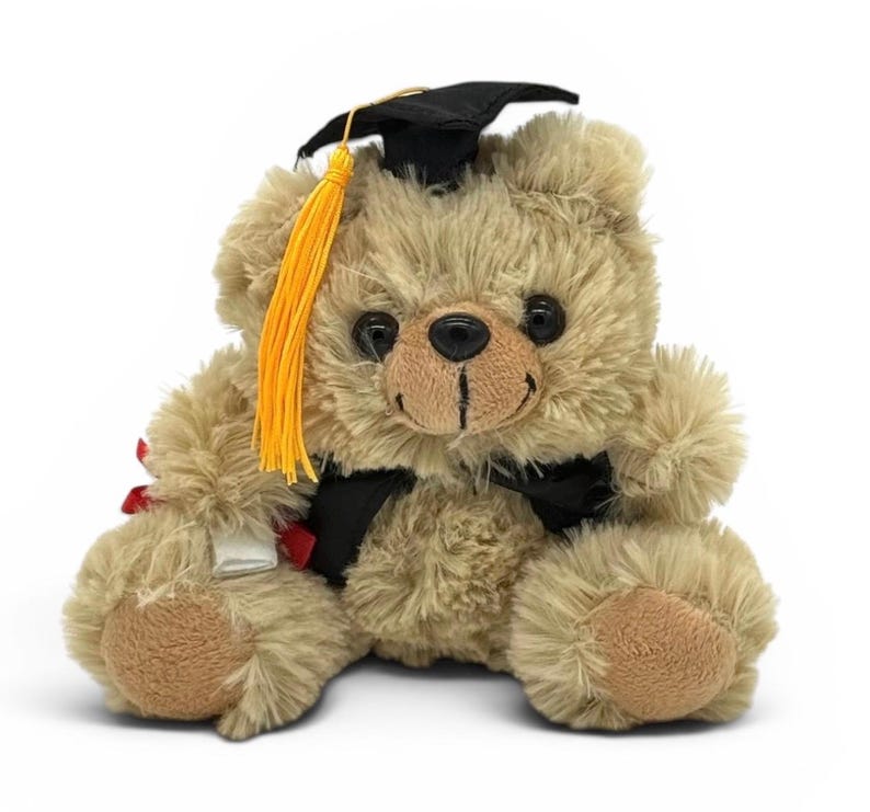 May include: A plush teddy bear in a graduation cap and gown. The bear is light brown with a black cap, a yellow tassel, and a black ribbon. It holds a small scroll, celebrating academic achievement.